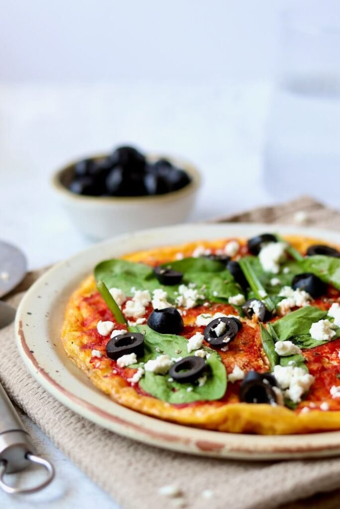 A pizza topped with spinach, black olives, and crumbled cheese sits on a plate, with a bowl of black olives and a pizza cutter nearby.