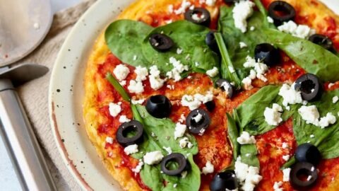 Delicious homemade pizza topped with spinach, black olives, and feta cheese on a rustic plate, served with a pizza cutter.