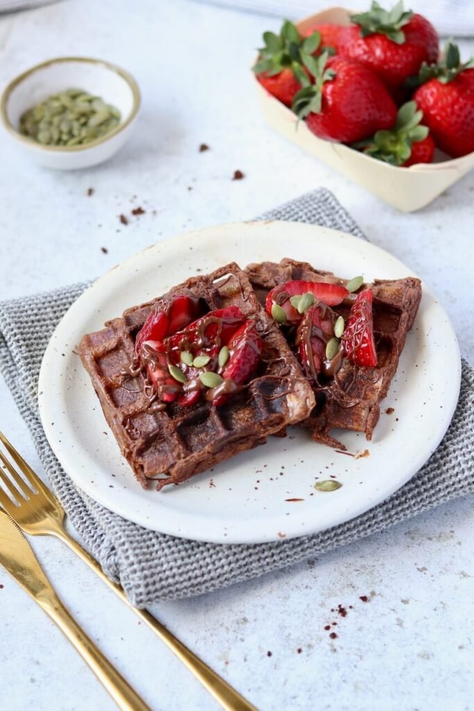Two chocolate protein waffles on a speckled ceramic plate, topped with sliced fresh strawberries, a drizzle of chocolate sauce, and pumpkin seeds. Styled on a grey waffle-knit cloth with gold cutlery alongside. A punnet of strawberries and a small bowl of pumpkin seeds sit in the background on a light marble surface.