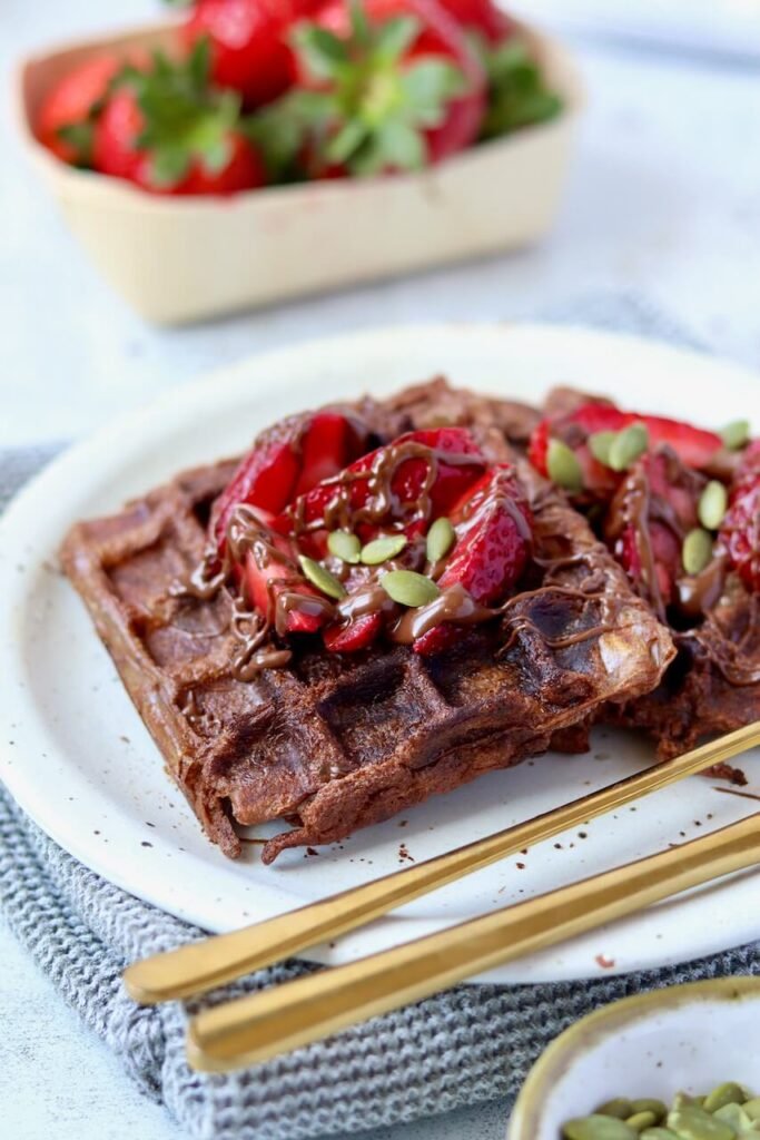 Close-up of two chocolate protein waffles on a speckled ceramic plate, topped with sliced fresh strawberries, a drizzle of chocolate sauce, and scattered pumpkin seeds. Gold cutlery rests on the plate. A punnet of strawberries is softly blurred in the background, with a small bowl of pumpkin seeds visible in the foreground corner, all styled on a grey waffle-knit cloth and light marble surface.