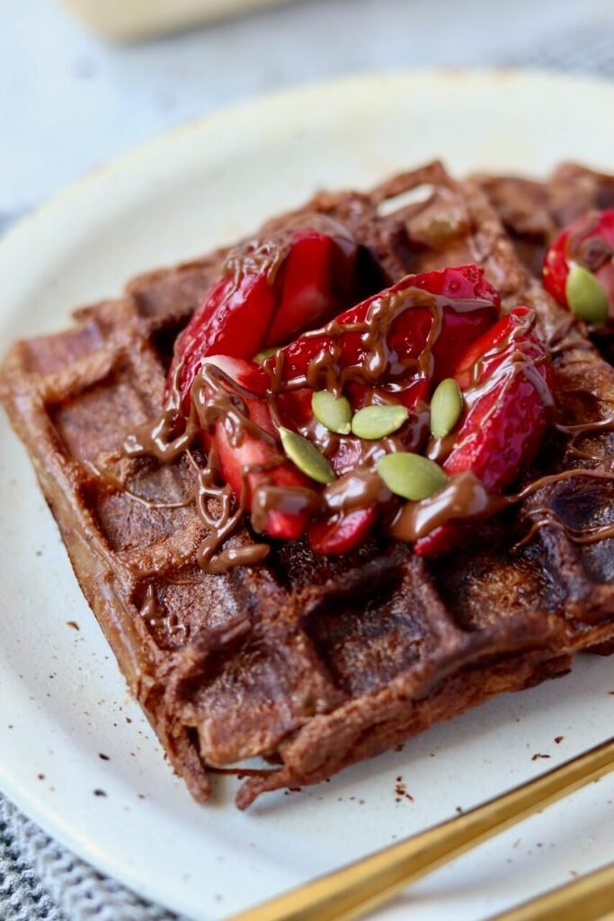 Extreme close-up of a chocolate protein waffle on a speckled ceramic plate, showing the golden-brown crispy grid texture in detail. The waffle is topped with juicy sliced strawberries, a generous drizzle of melted chocolate sauce, and pumpkin seeds. A gold fork is partially visible at the bottom right corner.