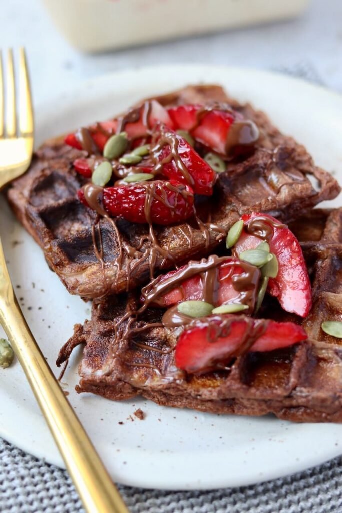 Two chocolate protein waffles stacked and overlapping on a speckled ceramic plate, each topped with chopped fresh strawberries, a drizzle of melted chocolate sauce, and pumpkin seeds. Gold fork and knife rest alongside the plate on a grey waffle-knit cloth, with a punnet of strawberries softly blurred in the background.