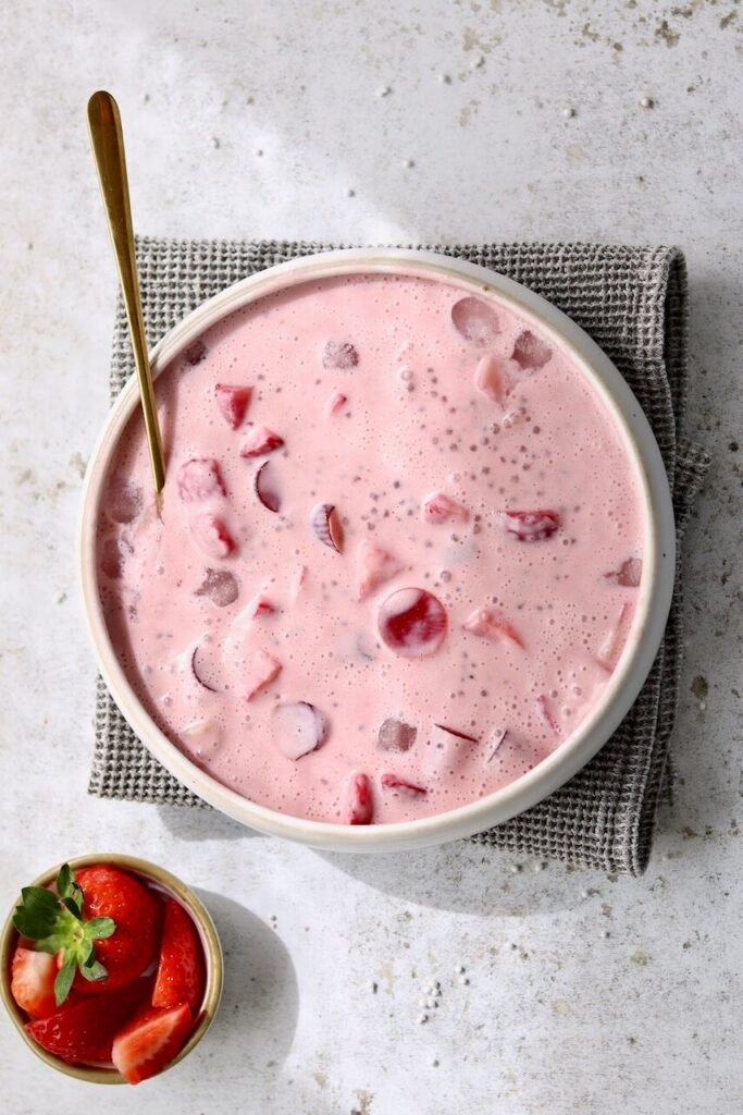 Overhead shot of a large white ceramic bowl filled with strawberries and cream sago pudding — a pale pink creamy mixture with visible sago pearls, chunks of strawberry, and rounded tapioca pieces — with a gold spoon resting inside. A small bowl of sliced fresh strawberries sits alongside on a light speckled surface with a grey waffle-knit cloth underneath the bowl.