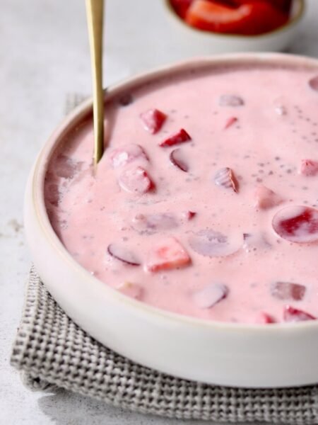 Close-up side angle of a white ceramic bowl filled with strawberries and cream sago pudding, showing the pale pink creamy texture with sago pearls and chunks of strawberry suspended throughout. A gold spoon is partially submerged in the pudding. The bowl rests on a grey waffle-knit cloth on a light speckled surface, with a small bowl of sliced strawberries softly blurred in the background.