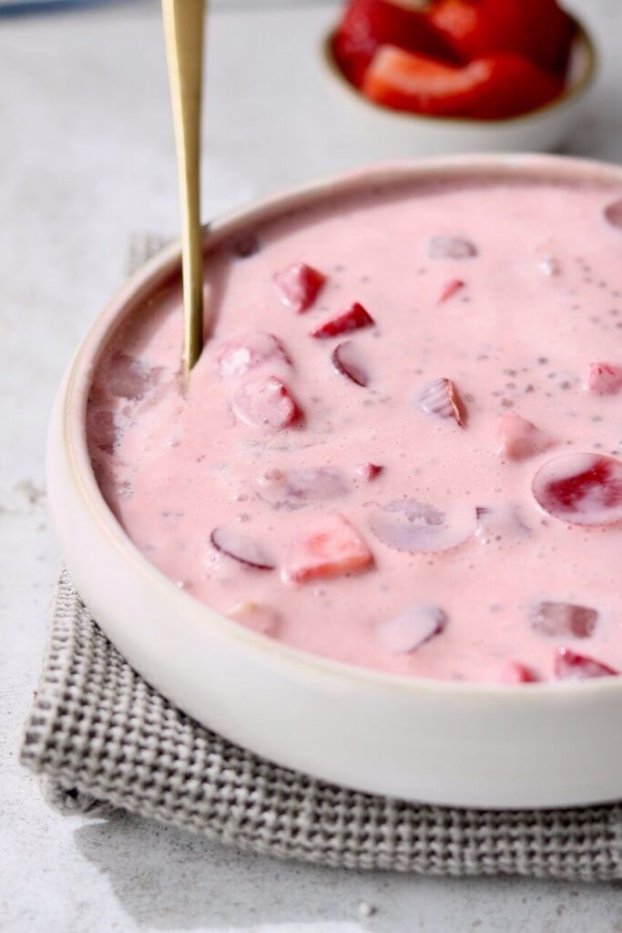 Close-up side angle of a white ceramic bowl filled with strawberries and cream sago pudding, showing the pale pink creamy texture with sago pearls and chunks of strawberry suspended throughout. A gold spoon is partially submerged in the pudding. The bowl rests on a grey waffle-knit cloth on a light speckled surface, with a small bowl of sliced strawberries softly blurred in the background.
