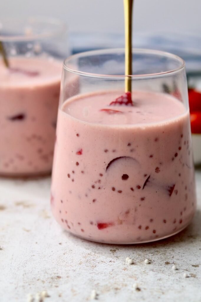 Close-up side angle of a white ceramic bowl filled with strawberries and cream sago pudding, showing the pale pink creamy texture with sago pearls and chunks of strawberry suspended throughout. A gold spoon is partially submerged in the pudding. The bowl rests on a grey waffle-knit cloth on a light speckled surface, with a small bowl of sliced strawberries softly blurred in the background.