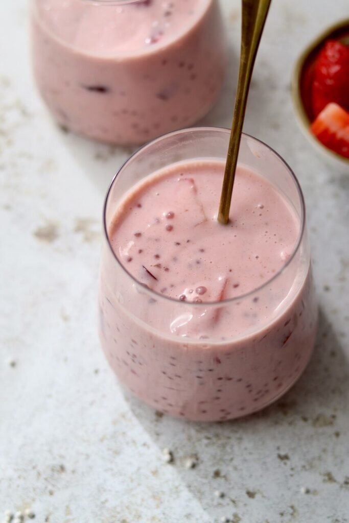 Close-up of two stemless glass tumblers filled with strawberries and cream sago, showing the pale pink creamy texture with sago pearls and pieces of strawberry, with a gold straw in the foreground glass. A small bowl of fresh strawberries is partially visible in the background on a light speckled surface.