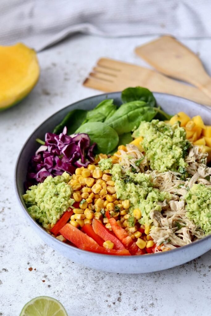 A vibrant high-protein shredded chicken salad bowl in a grey ceramic bowl, loaded with shredded chicken, sweetcorn, sliced red pepper, shredded red cabbage, baby spinach, diced mango, and a generous topping of chunky avocado chimichurri dressing. Styled on a light speckled surface with wooden salad servers, a halved mango, and a lime wedge in the background