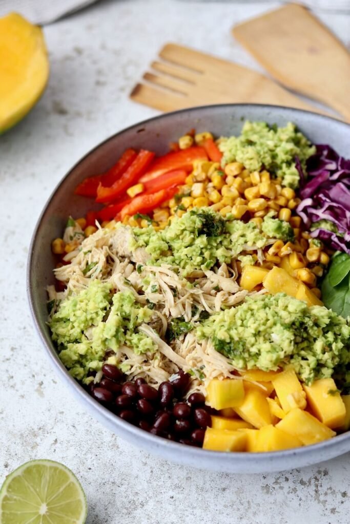 A colourful high-protein shredded chicken salad bowl in a grey ceramic bowl, featuring shredded chicken, black beans, diced mango, sweetcorn, sliced red pepper, shredded red cabbage, baby spinach, and a chunky avocado chimichurri dressing. Styled on a light speckled surface with wooden salad servers, a halved mango, and a squeezed lime wedge in the foreground.