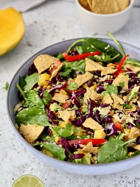 A tossed high-protein shredded chicken salad in a grey ceramic bowl, mixed together with baby spinach, rocket, shredded red cabbage, diced mango, sweetcorn, sliced red pepper, black beans, fresh coriander, and avocado chimichurri dressing, topped with broken protein corn chips. A small bowl of corn chips, a halved mango, and a squeezed lime wedge are styled around the bowl on a light speckled surface.