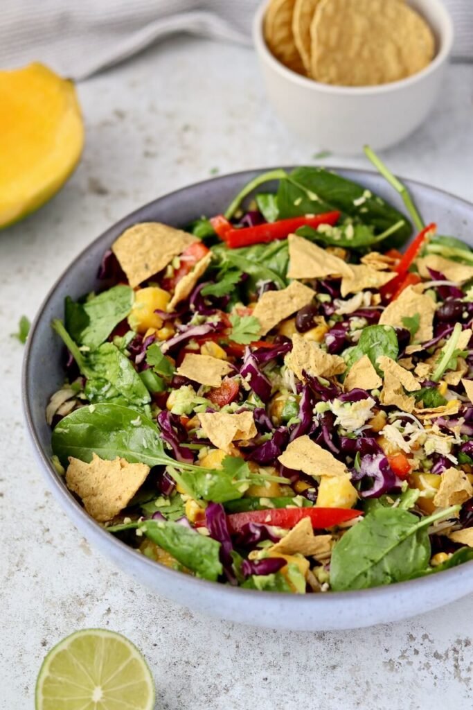 A tossed high-protein shredded chicken salad in a grey ceramic bowl, mixed together with baby spinach, rocket, shredded red cabbage, diced mango, sweetcorn, sliced red pepper, black beans, fresh coriander, and avocado chimichurri dressing, topped with broken protein corn chips. A small bowl of corn chips, a halved mango, and a squeezed lime wedge are styled around the bowl on a light speckled surface.