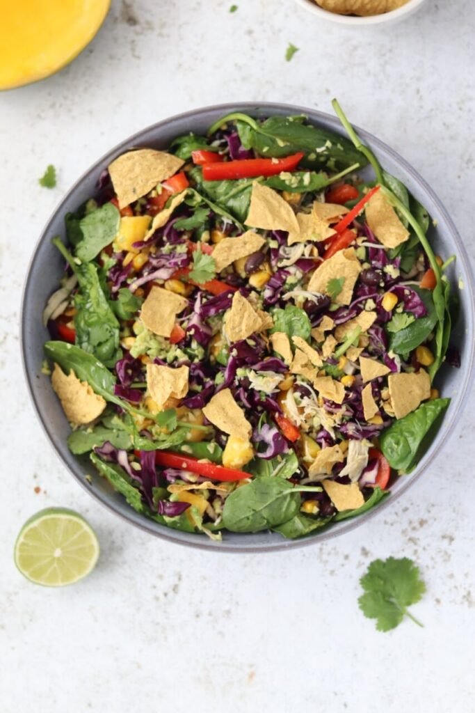 Overhead shot of a tossed high-protein shredded chicken salad in a grey ceramic bowl, packed with baby spinach, rocket, shredded red cabbage, diced mango, sweetcorn, sliced red pepper, black beans, shredded chicken, fresh coriander, and avocado chimichurri dressing, topped with broken protein corn chips. A squeezed lime half, fresh coriander leaves, a halved mango, and a small bowl of corn chips are scattered around the bowl on a light speckled surface.