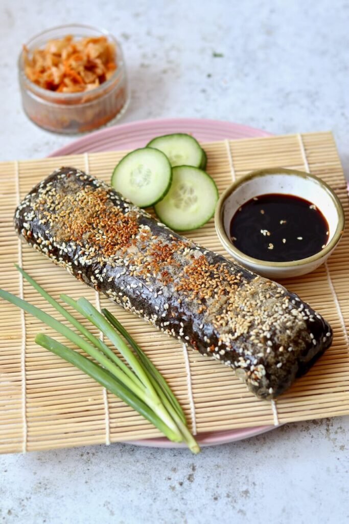 A whole rolled sushi sandwich wrap coated in sesame seeds, served on a pink plate atop a bamboo mat, surrounded by sliced cucumber, a small bowl of soy sauce, fresh spring onions, and a jar of kimchi.