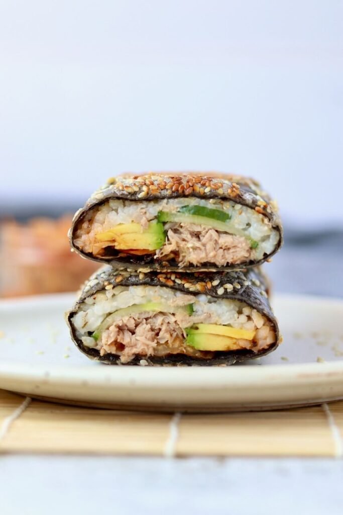 Close-up front view of a sliced sushi sandwich wrap stacked on a speckled ceramic plate, showing the cross-section layers of rice, tuna, avocado, and cucumber inside a sesame seed-coated nori wrap, with a jar of kimchi blurred in the background.