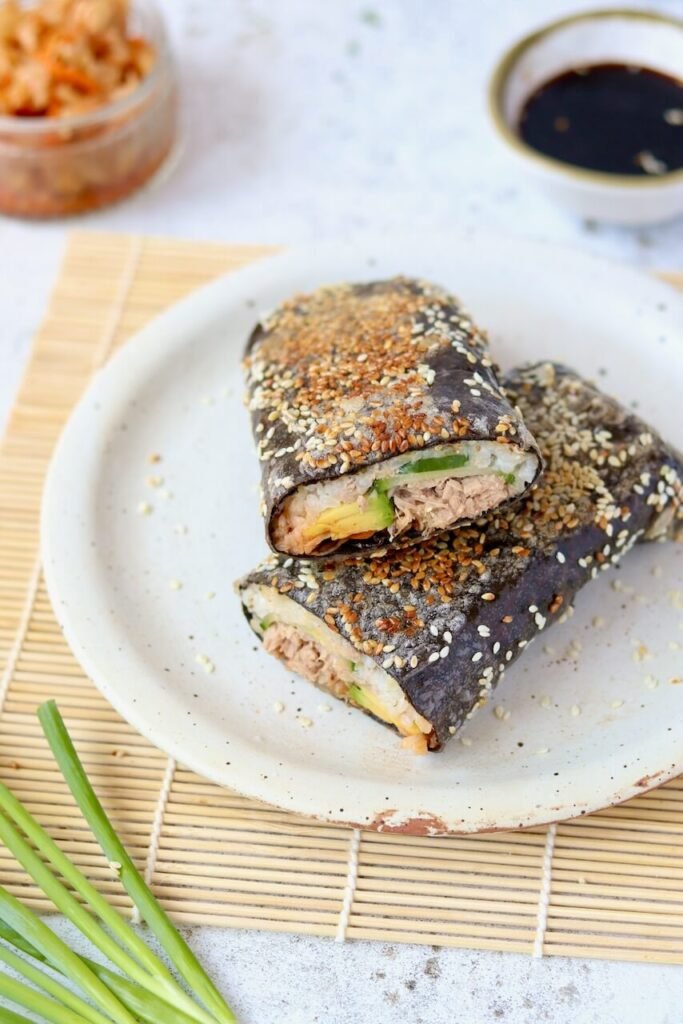 A sushi sandwich wrap coated in sesame seeds, cut into two halves and stacked on a speckled ceramic plate, revealing layers of rice, tuna, avocado, cucumber, and kimchi inside. Soy sauce, a jar of kimchi, and fresh spring onions are arranged around the plate on a bamboo mat