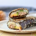 Close-up of a sushi sandwich wrap cut into two halves and stacked on a speckled ceramic plate, showing the cross-section of rice, tuna, avocado, and cucumber inside a sesame-coated nori wrap, with a jar of kimchi softly blurred in the background.