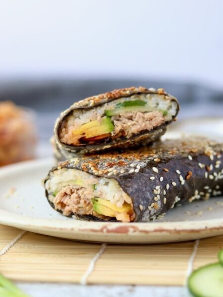 Close-up of a sushi sandwich wrap cut into two halves and stacked on a speckled ceramic plate, showing the cross-section of rice, tuna, avocado, and cucumber inside a sesame-coated nori wrap, with a jar of kimchi softly blurred in the background.