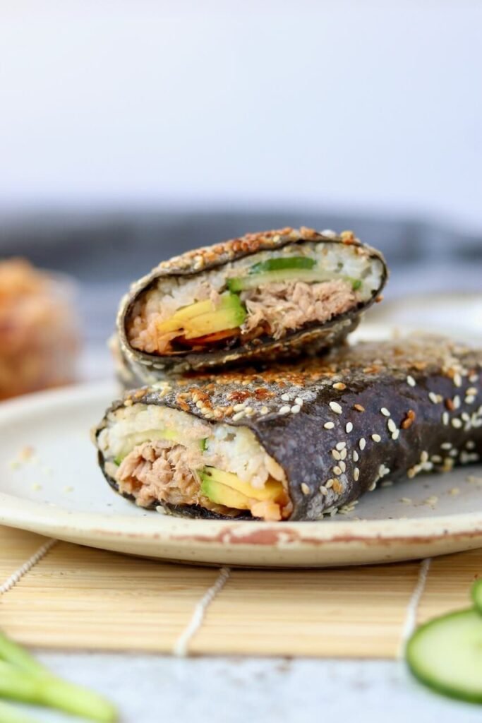 Close-up of a sushi sandwich wrap cut into two halves and stacked on a speckled ceramic plate, showing the cross-section of rice, tuna, avocado, and cucumber inside a sesame-coated nori wrap, with a jar of kimchi softly blurred in the background.