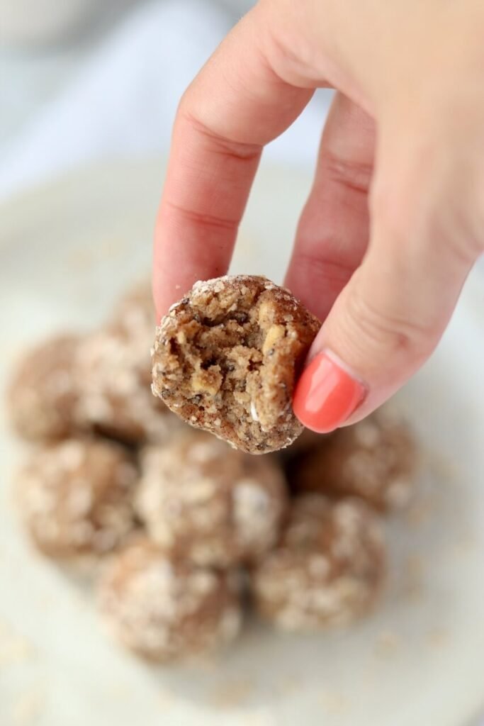 A hand holding a High Protein Tres Leches Cake Bite with a bite taken out, revealing the dense, chunky textured centre, held above a pile of more cake bites on a speckled ceramic plate.