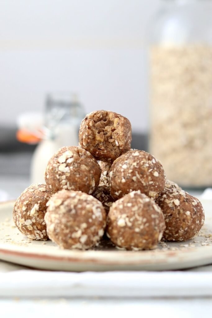 A pyramid stack of High Protein Tres Leches Cake Bites coated in oats on a speckled ceramic plate, with the top bite showing a bite taken out, photographed against a blurred background of a milk bottle and a jar of oats.
