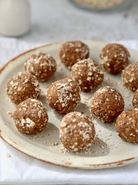 A pile of High Protein Tres Leches Cake Bites coated in rolled oats arranged on a speckled ceramic plate, with a glass of oat milk and a jar of oats softly blurred in the background on a white linen cloth.