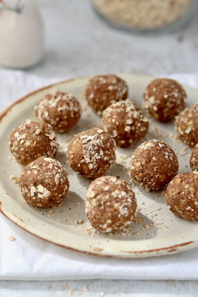 A pile of High Protein Tres Leches Cake Bites coated in rolled oats arranged on a speckled ceramic plate, with a glass of oat milk and a jar of oats softly blurred in the background on a white linen cloth.