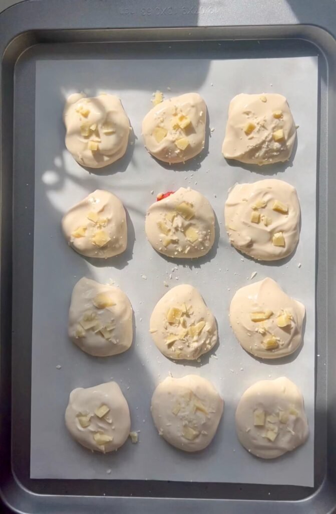 Twelve white chocolate-coated protein bites arranged in rows on a baking tray lined with parchment paper, topped with chopped white chocolate pieces, ready to set.