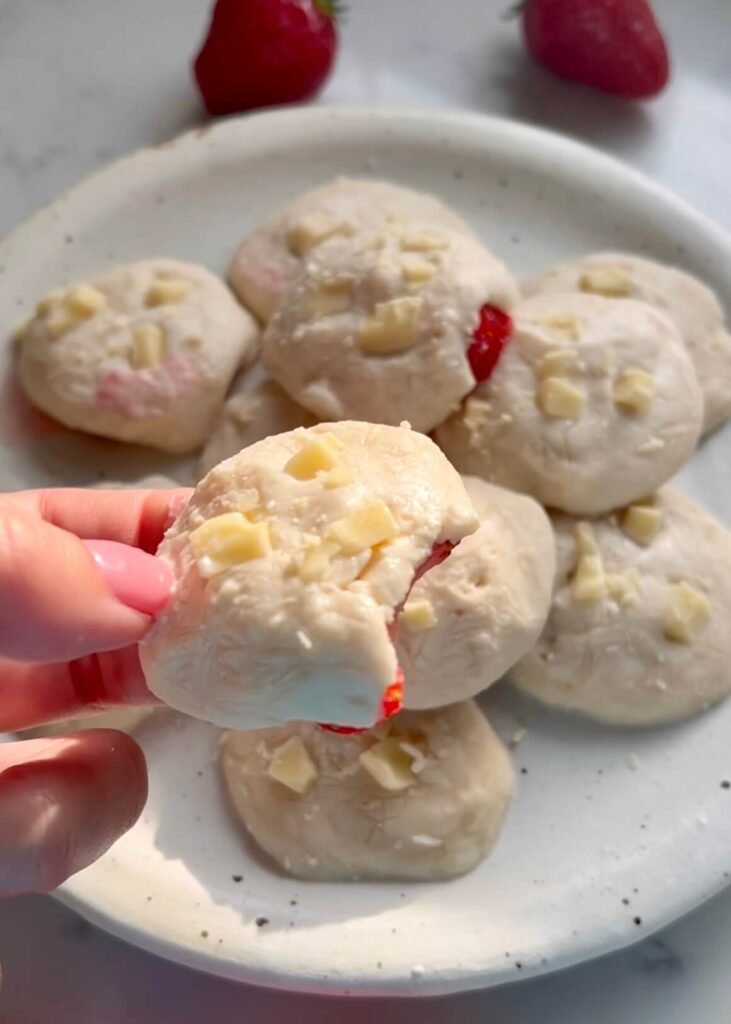 A hand holding a strawberry and white chocolate protein bite with a bite taken out, revealing a juicy strawberry centre and white filling, with whole bites piled on a speckled ceramic plate and fresh strawberries in the background.