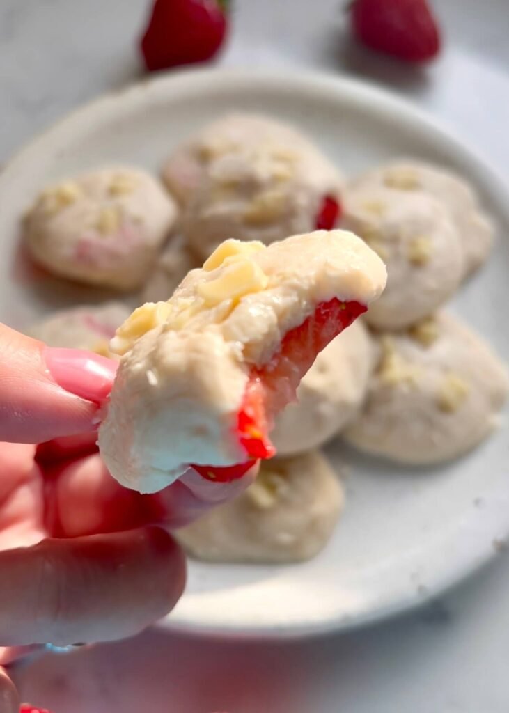 Close-up of a strawberry and white chocolate protein bite with a bite taken out, showing the layers of filling and whole strawberry inside, held in front of a plate of more bites and fresh strawberries.