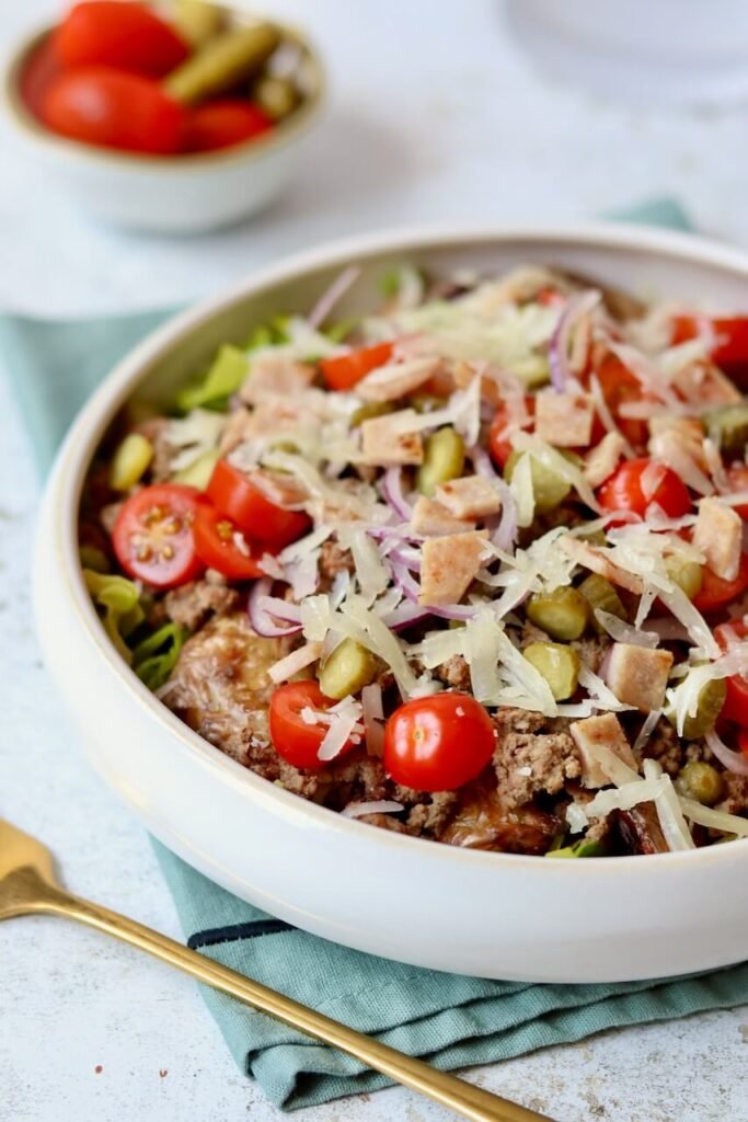 A white ceramic bowl filled with cheeseburger smashed potato salad — ground beef, smashed baby potatoes, cherry tomatoes, chopped gherkins, sliced red onion, shaved cheese, and crouton pieces — without the burger sauce, with a gold fork alongside on a teal linen cloth and a small bowl of tomatoes and gherkins in the background.