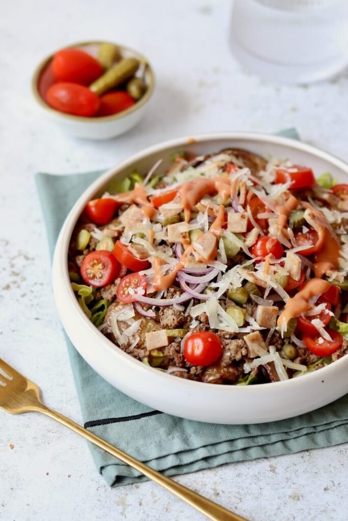 A high-protein cheeseburger smashed potato salad in a white ceramic bowl, loaded with seasoned ground beef, smashed baby potatoes, cherry tomatoes, chopped gherkins, sliced red onion, shaved cheese, and crouton-style bread pieces, drizzled with a pink burger sauce. A gold fork rests alongside on a teal linen cloth, with a small bowl of cherry tomatoes and gherkins blurred in the background.