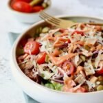 A white ceramic bowl of cheeseburger smashed potato salad topped with a generous drizzle of pink burger sauce, with a gold fork resting inside the bowl and a small bowl of cherry tomatoes and gherkins blurred in the background on a teal linen cloth.