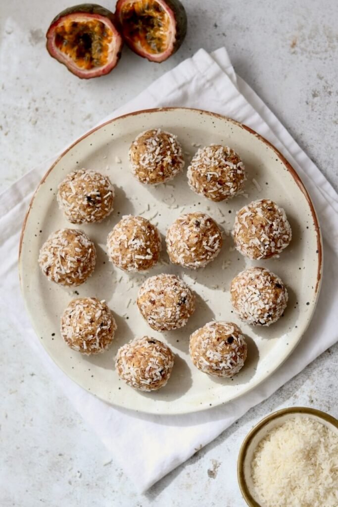 Overhead shot of twelve coconut-coated passion fruit protein bites arranged on a speckled ceramic plate on a white linen cloth, with a halved passion fruit in the top corner and a small bowl of desiccated coconut alongside.