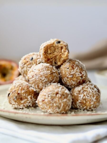 A pyramid stack of coconut-coated passion fruit protein bites on a speckled ceramic plate, with the top bite showing a bite taken out to reveal the dense oat and date interior, with a halved passion fruit visible in the background.