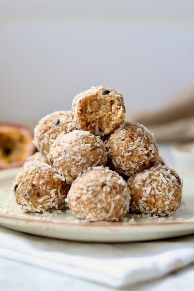 A pyramid stack of coconut-coated passion fruit protein bites on a speckled ceramic plate, with the top bite showing a bite taken out to reveal the dense oat and date interior, with a halved passion fruit visible in the background.
