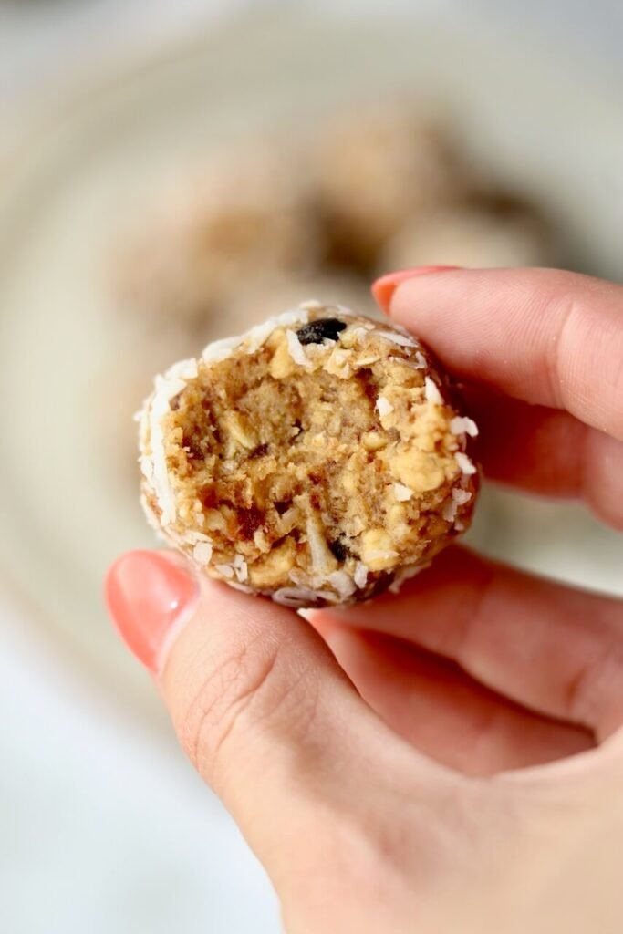 Close-up of a passion fruit protein bite with a bite taken out, held between two fingers, showing the chewy oat and date interior, with more bites blurred on a plate in the background.