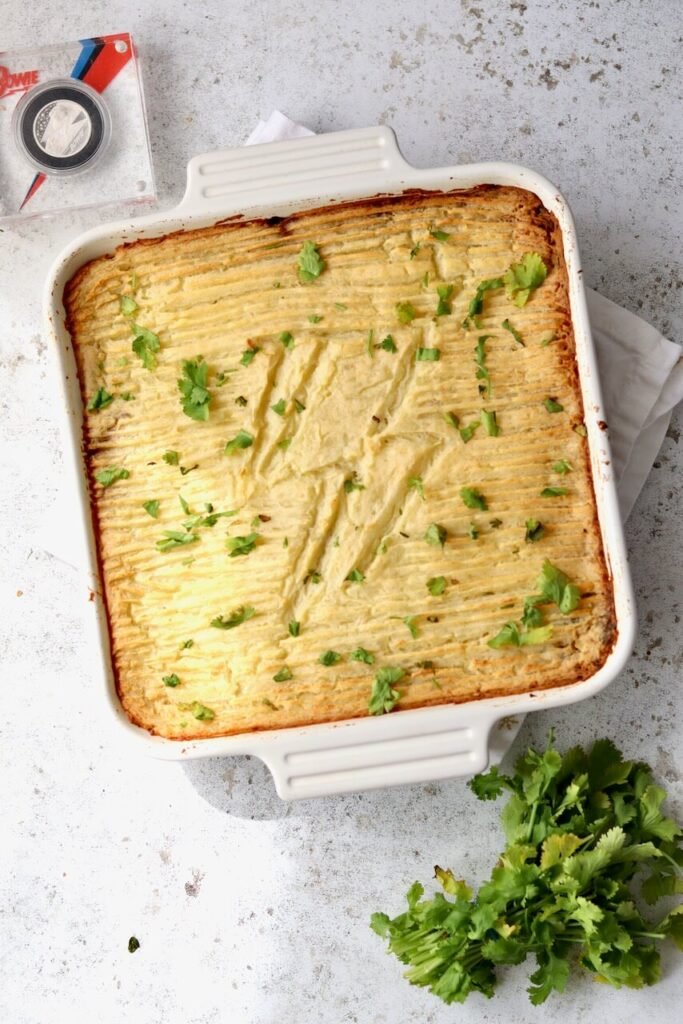 Overhead shot of a freshly baked high-protein cottage pie in a white ceramic baking dish, with golden fork-ridged mashed potato on top, lightly browned at the edges and garnished with fresh coriander leaves, styled on a light speckled surface.