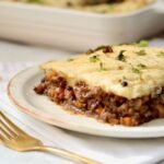 A side-on serving of high-protein shepherds pie on a speckled ceramic plate, showing the thick beef and lentil mince base topped with a golden mashed potato layer and scattered fresh coriander, with a gold fork alongside and the baking dish softly blurred in the background.