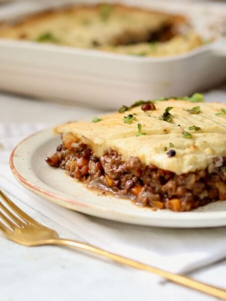 A side-on serving of high-protein shepherds pie on a speckled ceramic plate, showing the thick beef and lentil mince base topped with a golden mashed potato layer and scattered fresh coriander, with a gold fork alongside and the baking dish softly blurred in the background.