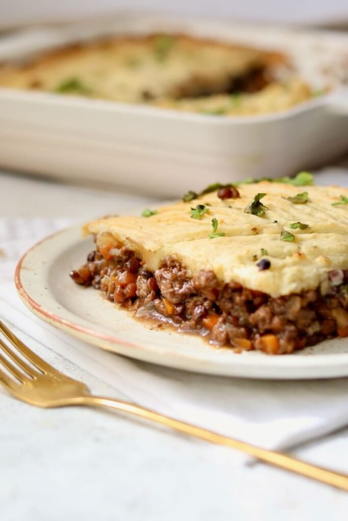 A side-on serving of high-protein shepherds pie on a speckled ceramic plate, showing the thick beef and lentil mince base topped with a golden mashed potato layer and scattered fresh coriander, with a gold fork alongside and the baking dish softly blurred in the background.