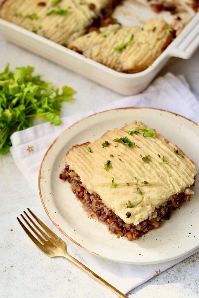 A serving of high-protein cottage pie on a speckled ceramic plate, showing a thick layer of seasoned beef and lentil mince topped with fork-ridged mashed potato and fresh coriander, with a gold fork alongside and the full baking dish visible in the background on a white linen cloth.
