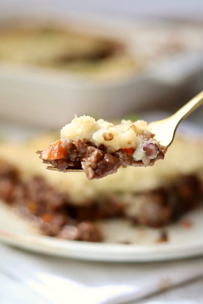 A gold fork holding a bite of high-protein cottage pie, showing the layers of mashed potato and beef and lentil mince with diced carrot, held in front of a blurred plate and baking dish.