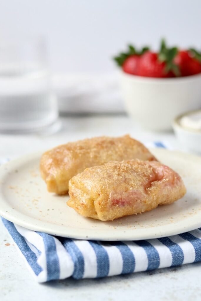 Two golden air-fried strawberry and cream cheese rice paper rolls on a speckled ceramic plate, dusted with cinnamon sugar, styled on a blue and white striped linen cloth with a bowl of fresh strawberries and a small dish softly blurred in the background.