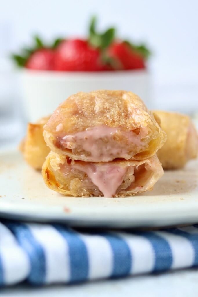 Close-up of two strawberry and cream cheese rice paper rolls stacked on a speckled ceramic plate, the top one sliced in half to show the creamy pink strawberry filling, with a bowl of fresh strawberries and a whole roll blurred in the background.