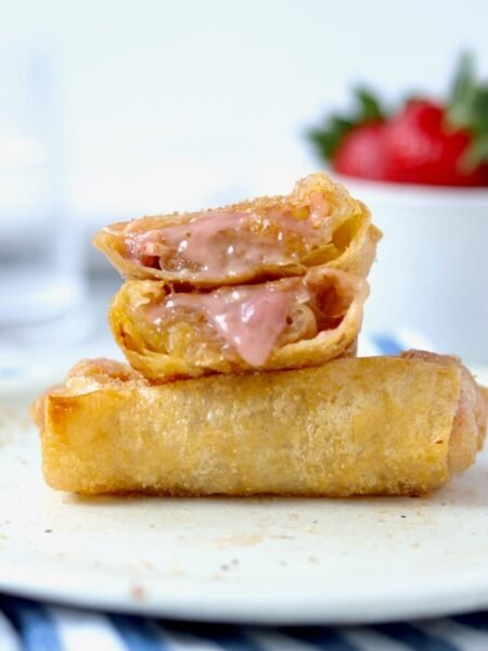 Two strawberry and cream cheese rice paper rolls stacked on a speckled ceramic plate, the top one sliced open to reveal the melted pink cream cheese and strawberry filling oozing out, with a bowl of fresh strawberries blurred in the background on a blue and white striped cloth.