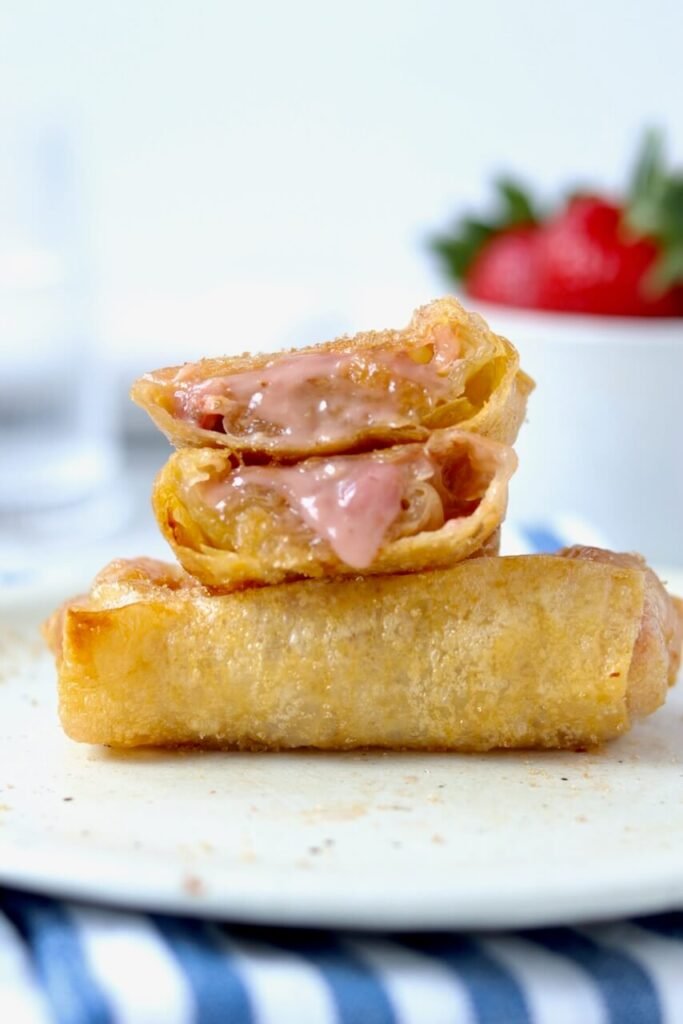 Two strawberry and cream cheese rice paper rolls stacked on a speckled ceramic plate, the top one sliced open to reveal the melted pink cream cheese and strawberry filling oozing out, with a bowl of fresh strawberries blurred in the background on a blue and white striped cloth.