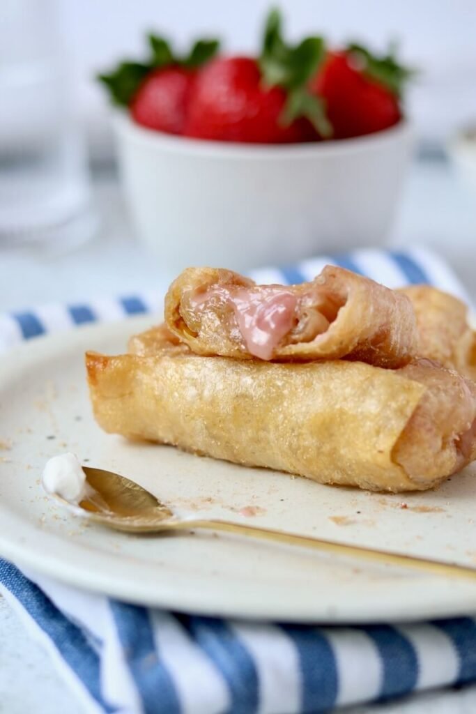 A strawberry and cream cheese rice paper roll broken open on a speckled ceramic plate, showing the melted pink cream cheese filling inside, with a whole roll beneath and a gold spoon alongside, with a bowl of fresh strawberries blurred in the background on a blue and white striped cloth.