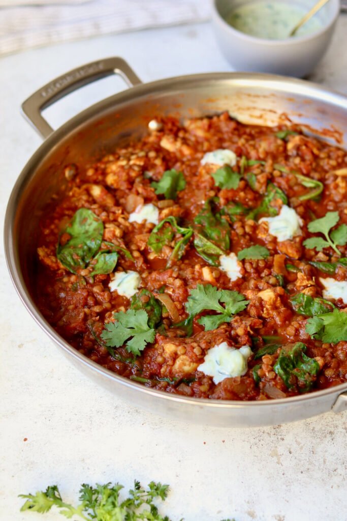 One-pot chicken dhansak simmering in a stainless steel pan, topped with wilted spinach, coriander and dollops of cucumber raita — an easy high-protein curry for weeknight meal prep.
