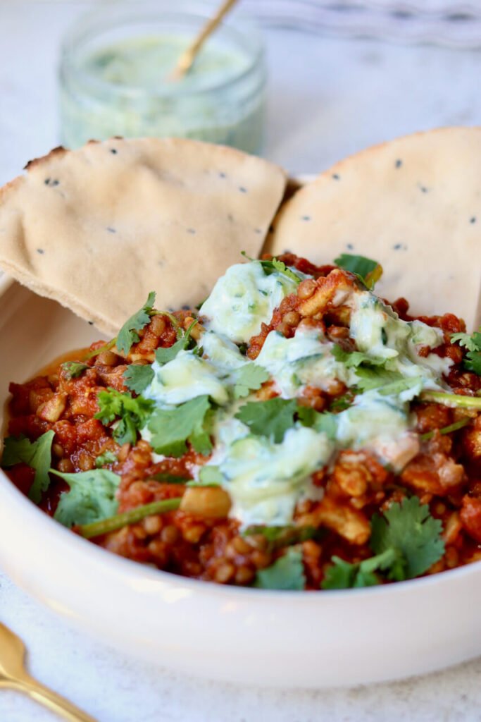 Bowl of lighter chicken dhansak with lentils, coriander and homemade cucumber raita, with seeded flatbread for scooping — an easy high-protein curry recipe.