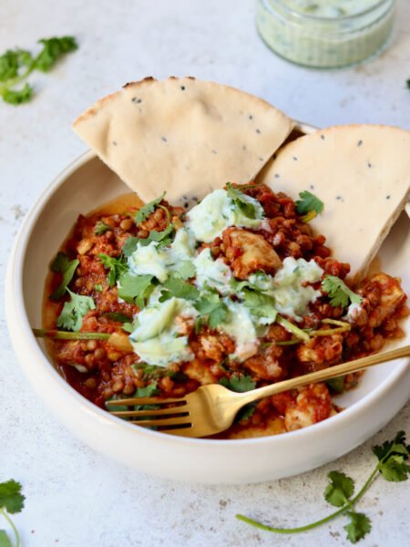 Healthy one-pot chicken dhansak served in a white bowl with lentils, fresh coriander, cucumber raita and seeded flatbread — a high-protein, lighter take on the fakeaway curry classic.